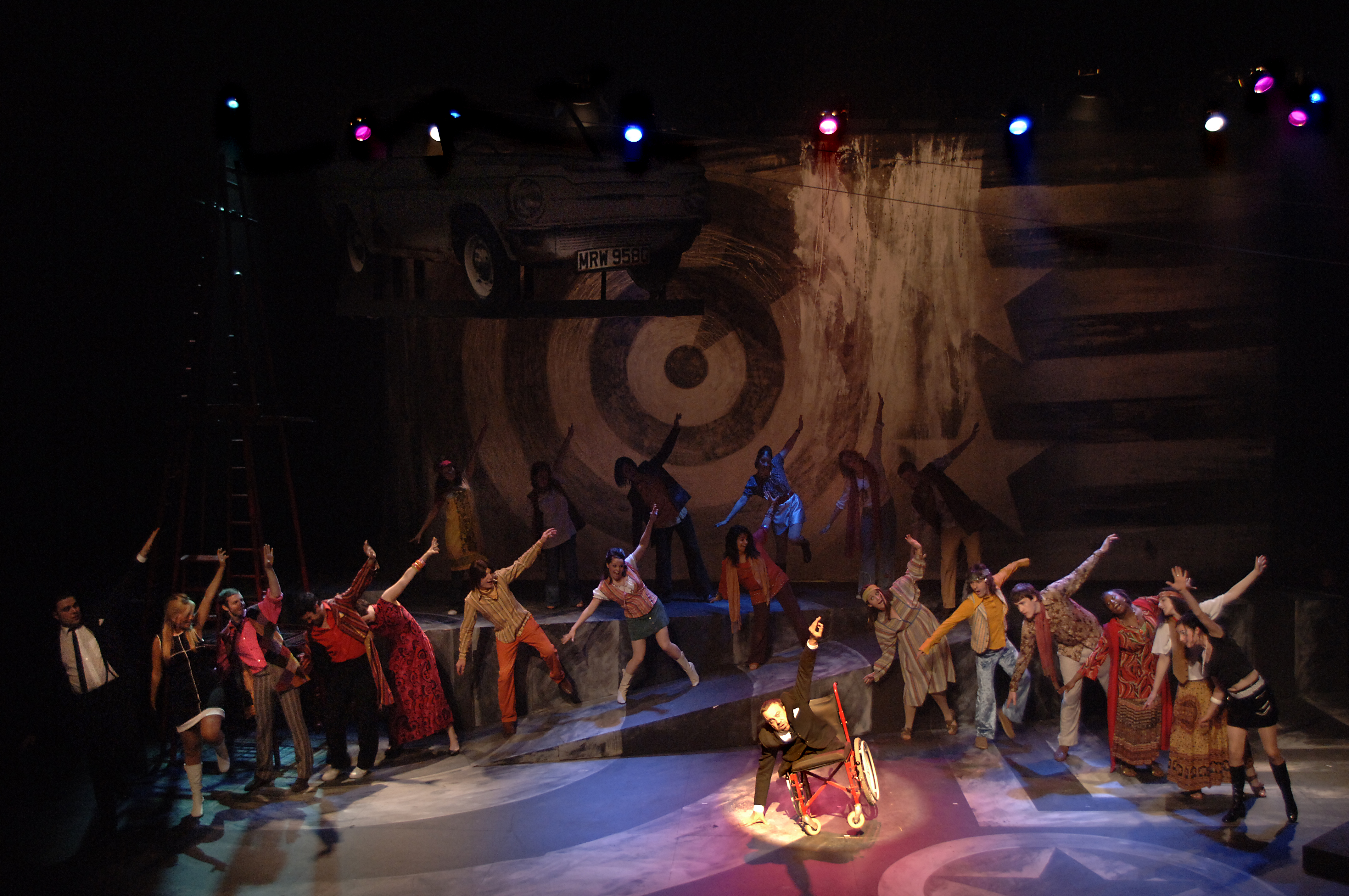 A man in a wheelchair leaning to one side with arms out like a plane with people in a circle behind copying