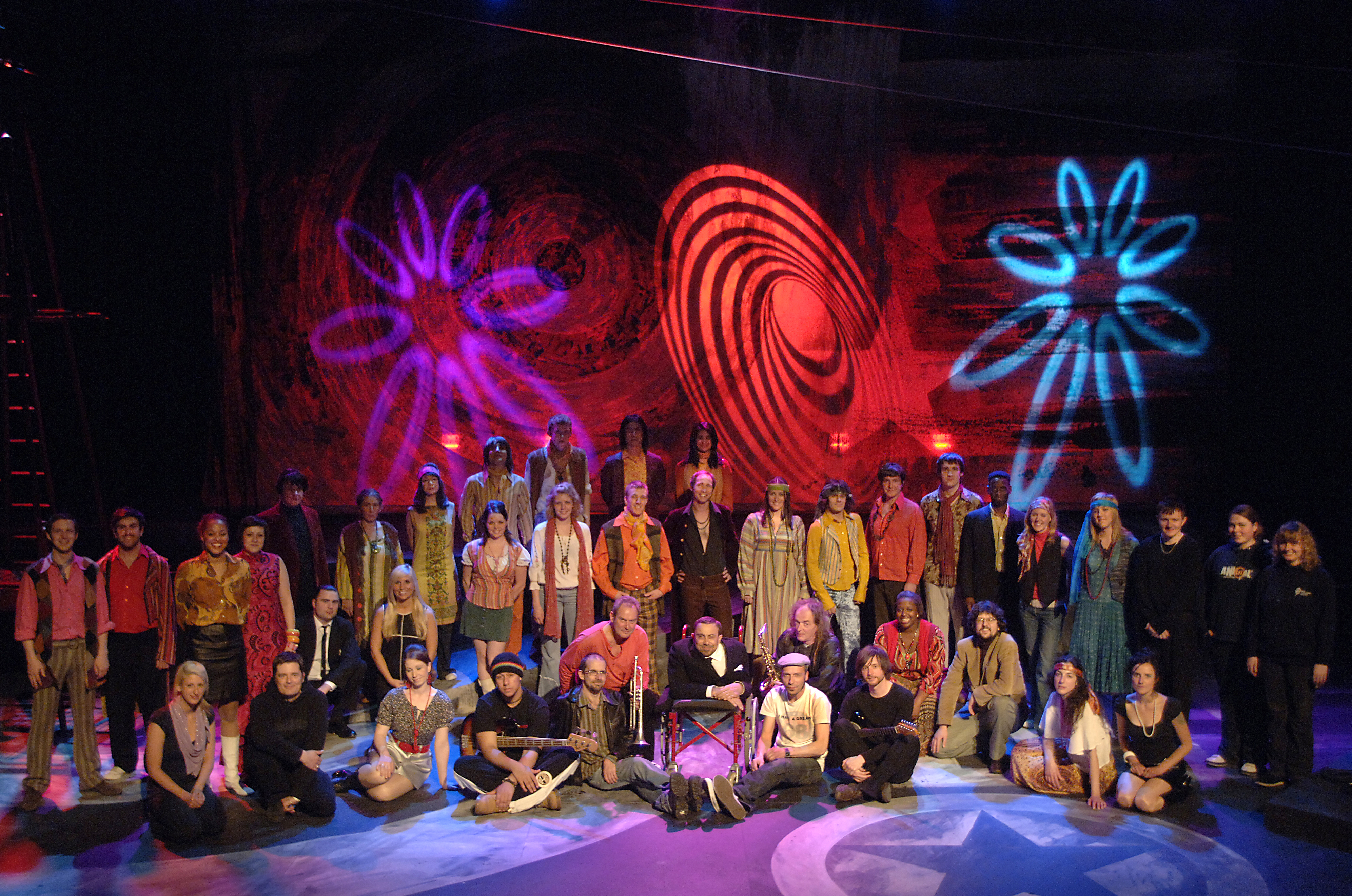A cast on stage in 70s period costume with psychedelic lights behind