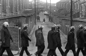 A woman walking up the street in between back to back terraces with police lining the street below in riot helmets.