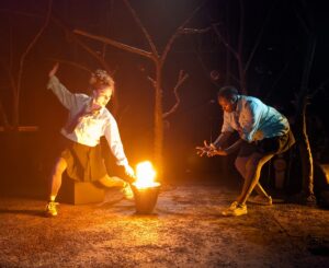 Two women dressed in school uniform moving around a fire in a bucket in a theatre set forest
