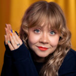Daisy Ann in a thick black jumper smiling with palms together next to her face. 