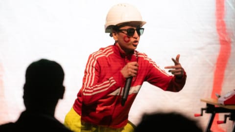 A woman in construction wear and glasses, rapping into a microphone