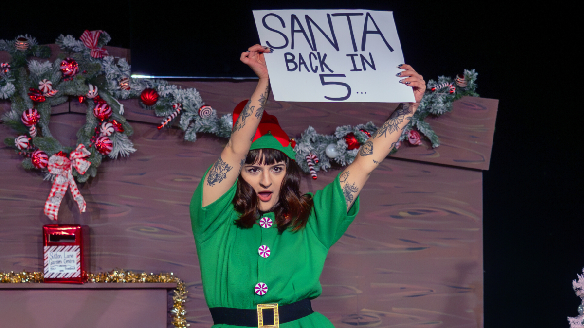 A woman dressed as an elf holding a sign that says Santa Back in 5 mins