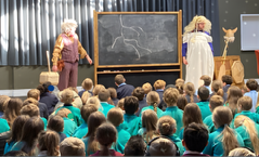 Two loopy male professors in drag with a blackboard in front of hundreds of school children