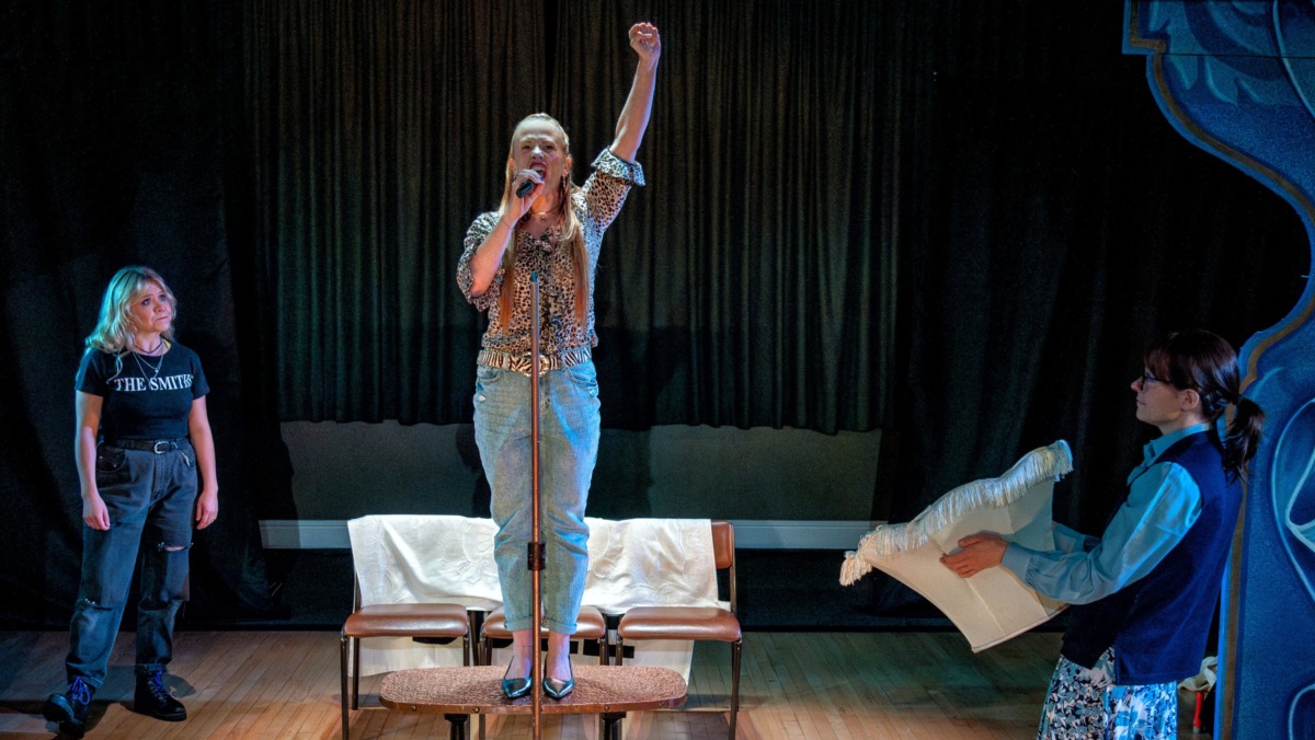 A woman on a coffee table making a pretend speech whilst her sister using a lampshade as a spotlight